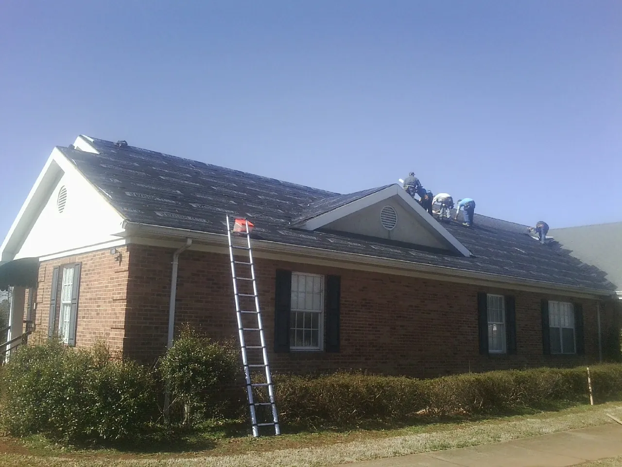 Crew tearing shingles off a brick ranch roof with a ladder set — Metal Roof Repair work by our crew in Wyocena