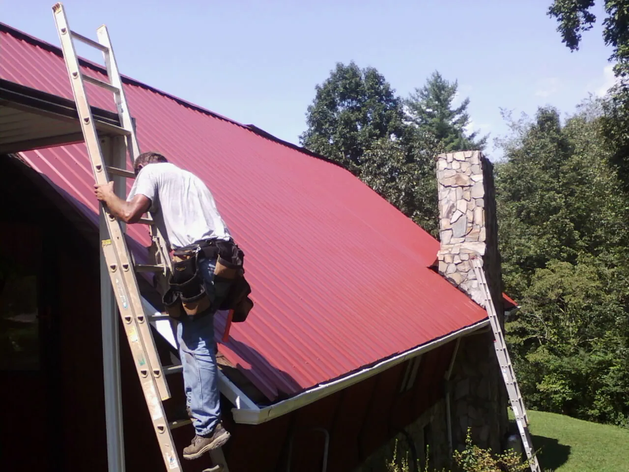 Roofer on a ladder beside a red metal roof and stone chimney — Soffit & Fascia Repair by Roofing Brothers in Wyocena